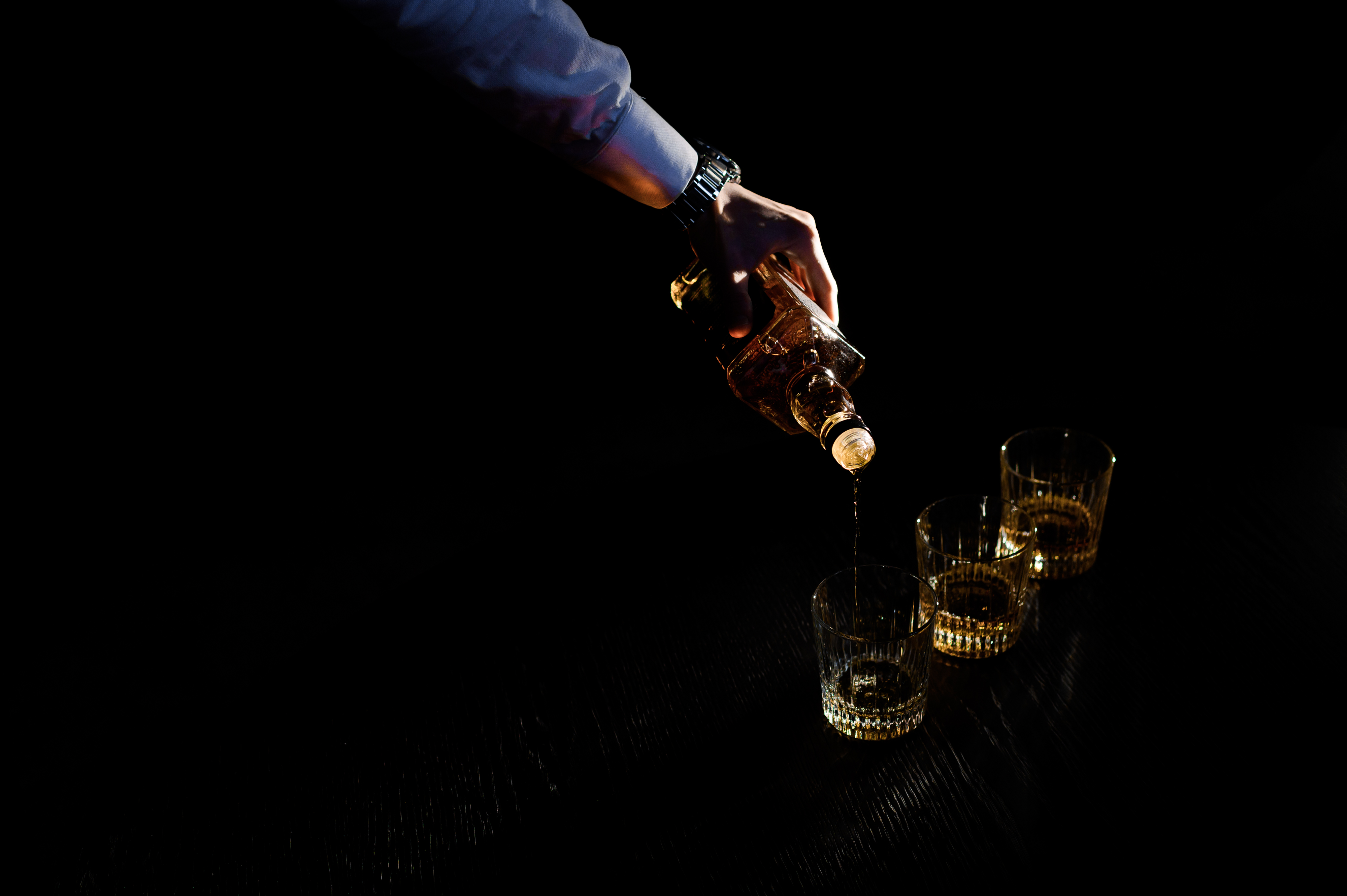  close-up-hand-holding-bottle-pours-drink-into-glass-black-dark-space-concept-celebrating-stag-party-barman-cocktails
